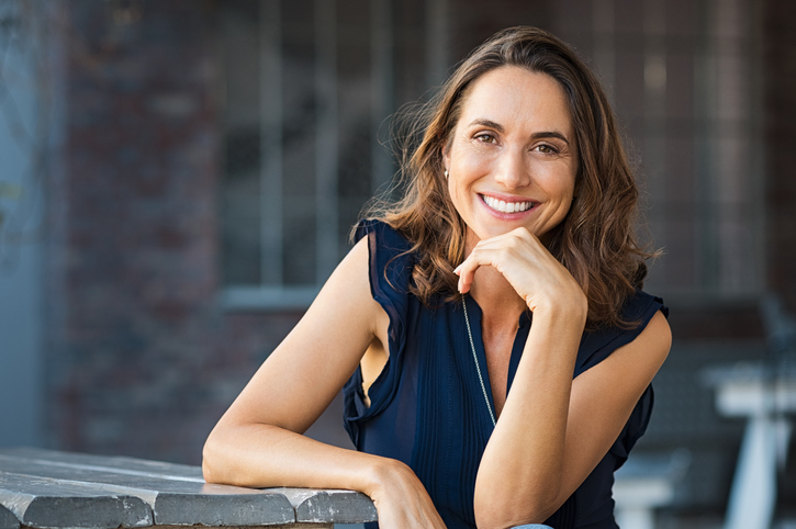 Portrait of beautiful mature woman sitting at coffee shop. Happy hispanic smiling woman sitting on a bench in outdoor cafeteria looking at camera. Portrait of carefree woman relaxing on bench.