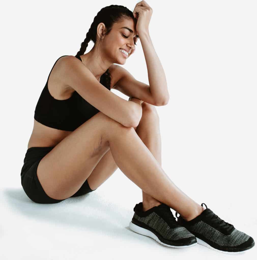 Woman sitting on floor on white background wearing athletic gear with spider veins on her legs