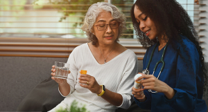 A young nurse or caregiver in scrubs showing a medicine bottle to an older female patient