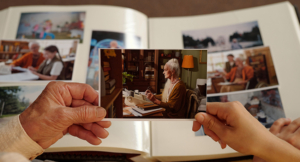 Senior Caucasian woman holding photograph of herself at sewing machine while sitting with child, both looking at photo album filled with family memories and past moments together