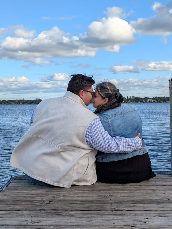 Jose and Rosie Garza Lopez are sitting on the edge of a dock near the water, hugging each other.