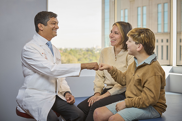Manish N. Shah greets a child patient with a fist bump while the child sits beside their parent in a bright clinic exam room with large windows.