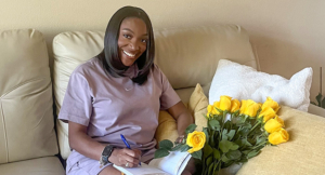 Prestal sitting on the couch, holding a pen and a journal next to a bouquet of yellow roses
