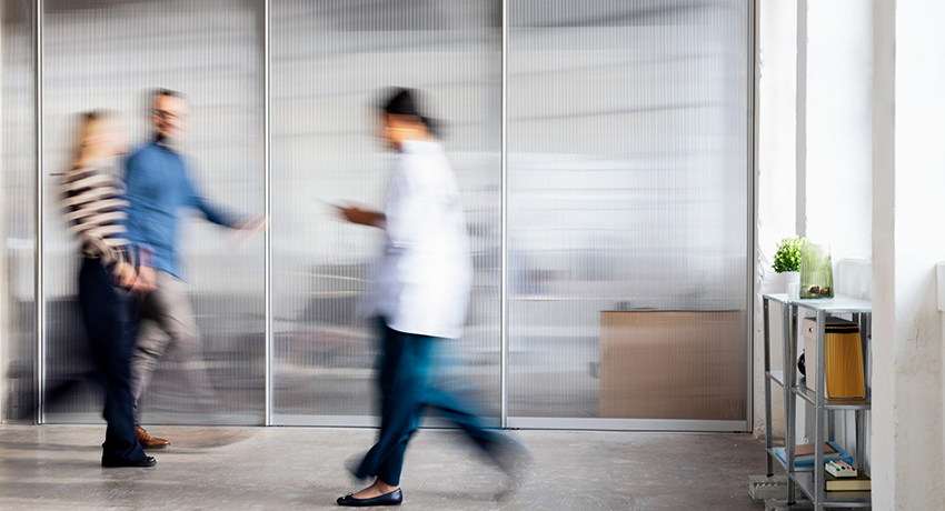 Blurred medical staff and visitors walk through a bright clinic corridor, conveying motion and busy healthcare.