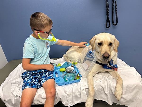 The boy sits on an exam room bed with Boris the dog and uses a toy stethoscope on Boris. 